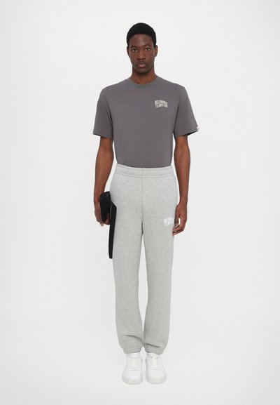 Grey cotton t-shirt with logo on chest, paired with grey sweatpants featuring an elastic waistband and logo print. White sneakers, holding a black bag.