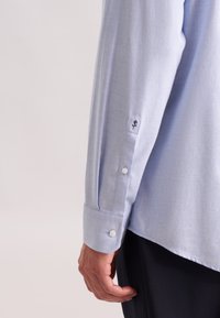 Camicetta a maniche lunghe azzurro chiaro realizzata con un tessuto a trama liscia. Presenta polsini abbottonati e un piccolo emblema ricamato sulla manica.