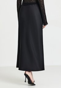 Donna che indossa una lunga gonna nera in satin, un top in pizzo nero e scarpe slingback nere con tacco alto, in piedi contro uno sfondo bianco.
