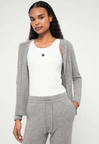 Cardigã cinza de malha sobre uma t-shirt branca com um logo de urso preto. O modelo está a usar calças de treino cinzas combinando, com cintura elástica e cordão.