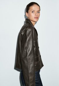 Una donna con capelli lisci raccolti indossa una giacca di pelle marrone scuro e jeans scuri, in piedi di lato contro uno sfondo chiaro e semplice.
