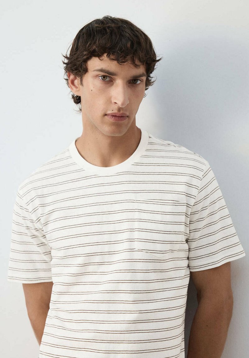 Joven con cabello corto, rizado y oscuro, vistiendo una camiseta blanca con finas rayas horizontales negras, de pie contra un fondo claro y liso.