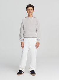 Jasnoszara dzianinowa bluza z kapturem o szerokim dekolcie, zestawiona z białymi spodniami i czarnymi tenisówkami z beżową podeszwą.