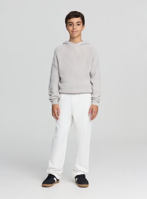 Felpa con cappuccio in maglia grigio chiaro con scollo ampio, abbinata a pantaloni bianchi e sneakers nere con suola color cuoio.