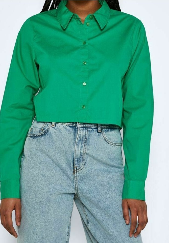Personne portant une chemise courte boutonnée vert vif et un jean taille haute bleu clair, les mains détendues le long du corps.
