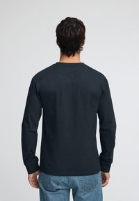 Svart T-shirt med lång ärm i bomull, med rund halsringning och rak skärning. Bärs med blå jeans, visar bakifrån.