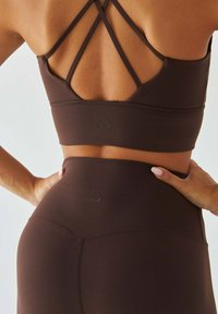 Brązowy sportowy biustonosz z krzyżującym się paskiem. Charakteryzuje się gładką teksturą i wytłoczonym logo serca na plecach oraz na pasie dopasowanych legginsów.