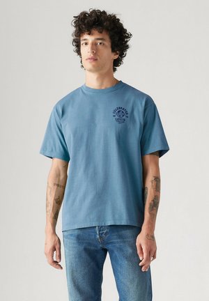 Giovane uomo con capelli ricci indossa una t-shirt blu Levi's e jeans blu, mostrando diversi tatuaggi su entrambe le braccia.