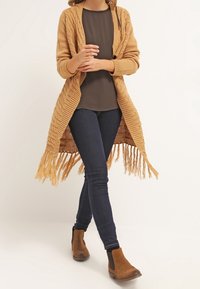 Beige neulecardigan, jossa on tekstuuroitu kuvio, avoin etuosa ja tupsudetailit. Päällä tumman paidan kanssa, yhdistetty sinisiin farkkuihin ja ruskeisiin nilkkureihin.