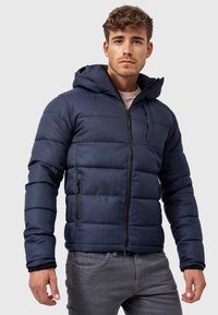 Marineblå puffjakke med hætte, der har en lynlås foran, sidelommer og et kviltet design med en let struktureret overflade.