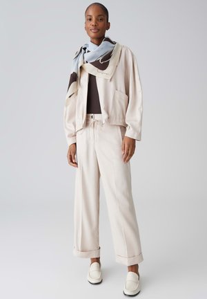 Donna in piedi che indossa una giacca beige e pantaloni a gamba larga, top nero, sciarpa fantasia e mocassini bianchi su uno sfondo neutro.