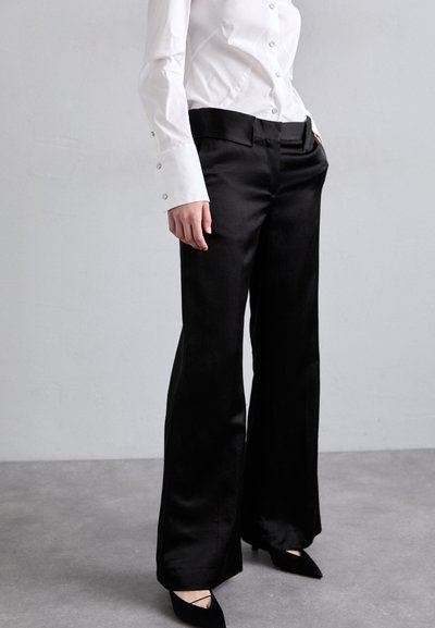 Pantaloni larghi in satin nero con una texture liscia e una vestibilità su misura, abbinati a una camicia bianca con bottoni lucenti e maniche lunghe.