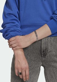 Modrá mikina s ohrnutými rukávy, šedé džínové kalhoty, stříbrný náramek na zápěstí a černý kámen na prstu. Viditelné jemné textury.