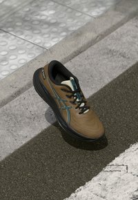 Zapatilla deportiva marrón con acentos negros y en tonos verde azulado, que presenta una parte superior texturizada, un sistema de cordones y una suela gruesa y contorneada.