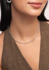 Collier en chaîne argentée avec des maillons ovales entrelacés, porté sur un haut en tissu tan. La texture lisse et la finition polie mettent en valeur le design.