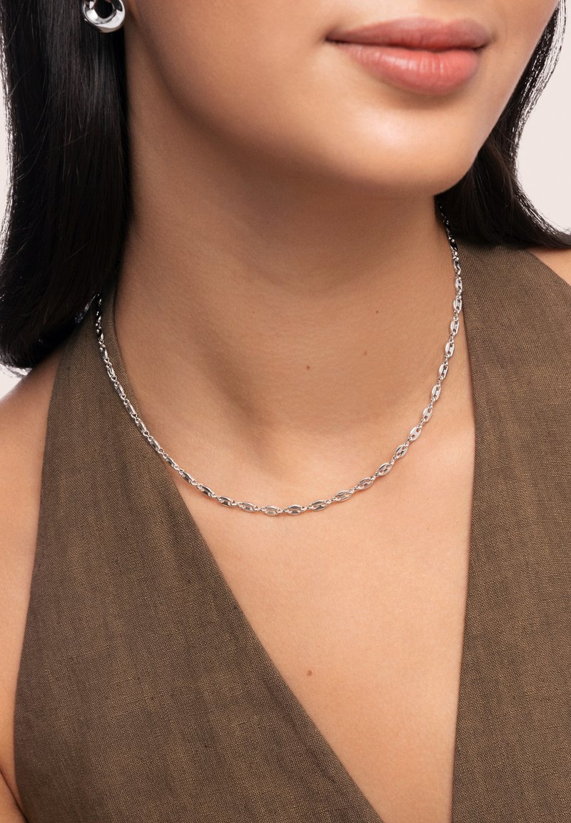 Collier en chaîne argentée avec des maillons ovales entrelacés, porté sur un haut en tissu tan. La texture lisse et la finition polie mettent en valeur le design.