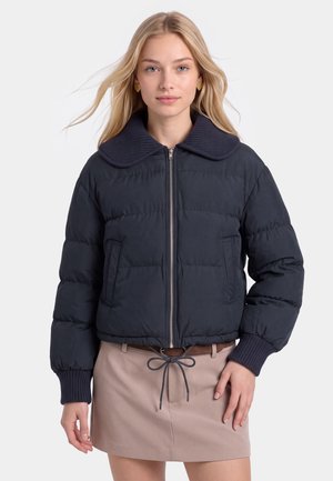 Jeune femme aux cheveux blonds portant une veste matelassée noire avec un large col et une jupe beige avec une ceinture marron et un cordon.