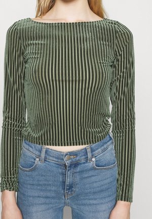 Femme portant un crop top en velours vert à manches longues avec des rayures verticales, associé à un jean bleu taille haute.