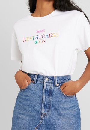 Kobieta nosząca białą koszulkę Levi Strauss z wielokolorowym logo i wysokim stanem niebieskie dżinsy, ręce w kieszeniach.