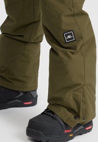 Gröna tekniska snöbyxor med en svart logopatch vid knät, utrustade med förstärkta knäpaneler och en lös passform över svarta snöskor.