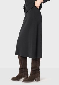 Jupe noire mi-longue en tissu lisse, associée à des bottes en daim marron jusqu'au genou avec un talon bloc.