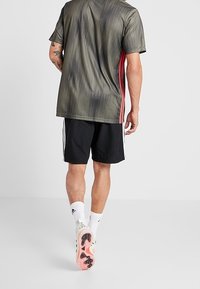 Uomo che indossa pantaloni corti neri, una maglietta sportiva grigia e rossa, calze bianche e scarpe da calcio bianche mentre cammina su uno sfondo grigio chiaro uniforme.