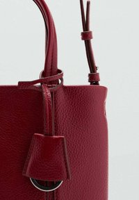 Rode leren handtas met een gestructureerde afwerking. Voorzien van twee handvaten, een decoratief accent en een strak ontwerp met een cirkelvormig sierbeslag.