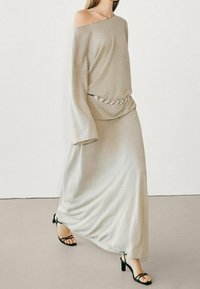 Beiges, schulterfreies Maxikleid mit langen Ärmeln und gerafftem Taillenbereich, kombiniert mit schwarzen Riemchen-High Heels. Der strukturierte Stoff fällt elegant.