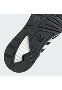 Suela de zapatilla en blanco y negro que muestra un caucho texturizado con profundas ranuras, un logo de adidas y una sección de media suela en blanco que contrasta.