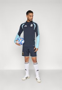 Farda de futebol masculino com uma parte superior de mangas longas azul-marinho com detalhes e logótipo em azul claro, calções combinando, meias brancas e botas de futebol.