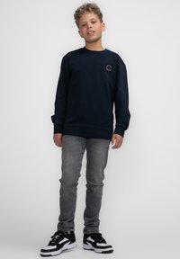 Marineblauwe sweatshirt met lange mouwen, ronde hals, en een cirkelvormige logo patch op de borst, gedragen met lichtgrijze slim-fit jeans en zwarte sneakers.