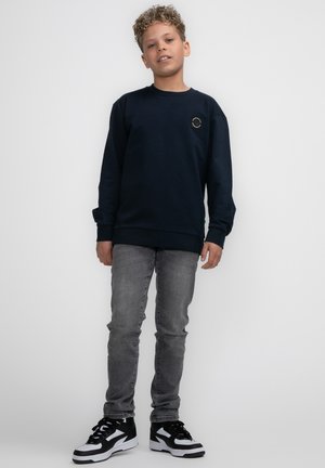 Felpa blu navy con maniche lunghe, colletto a girocollo e una patch circolare con logo sul petto, abbinata a jeans slim fit grigio chiaro e sneakers nere.