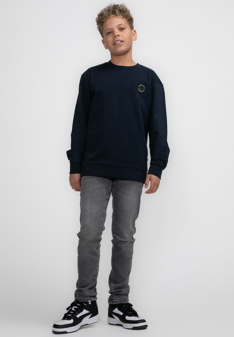 Marineblauwe sweatshirt met lange mouwen, ronde hals, en een cirkelvormige logo patch op de borst, gedragen met lichtgrijze slim-fit jeans en zwarte sneakers.