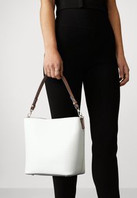 Vit läderhinkväska med en slät textur, med en brun justerbar rem, hålls av en hand, i kombination med svarta leggings.