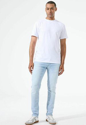 Man standing wearing a plain white short-sleeve t-shirt, light blue jeans, and white sneakers against a white background.
