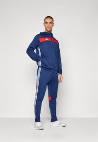 Navy tracksuit with red and white accents. Hoodie features drawstrings; pants have side stripes and a red patch. Material appears smooth.