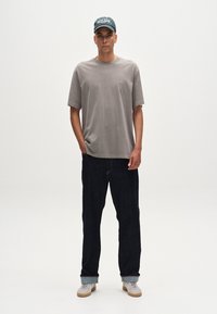 Tricou din bumbac gri, cu mâneci scurte, purtat cu blugi negri. Accesoriile includ o șapcă verde și adidași de culoare deschisă.