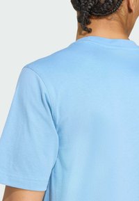 Světle modré bavlněné tričko s krátkým rukávem, se zdobným výstřihem do kulata a hladkou texturou. Viditelné prošívání poblíž švu na rameni.