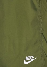 Olivgröna tygshorts med vit Nike-logotyp i nedre vänstra hörnet. Materialet ser slätt ut med synliga sömmar längs sömmarna.