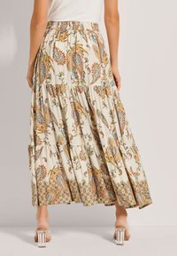 Frau in einem cremefarbenen, gestuften Maxirock mit Paisley- und Blumenmustern, kombiniert mit Sandalen mit transparenten Absätzen, die vor einem schlichten Hintergrund steht.