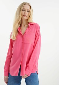 Růžová košile s dlouhým rukávem, zapínáním na knoflíky a kapsou, vyrobená z lehkého materiálu, s volným střihem a bočními rozparky, kombinovaná s modrými džíny.