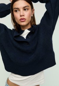 Navy blue cropped sweater in fuzzy texture with a shawl collar, short sleeves, and three black buttons at the neck, layered over a white top.
