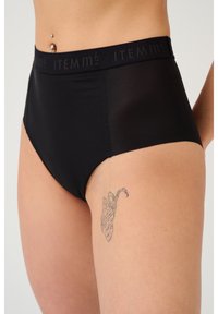 Mutande nere a vita alta con una texture liscia, dotate di un'ampia fascia elastica decorata con "ITEM m6." Mostrano un tatuaggio sulla coscia.