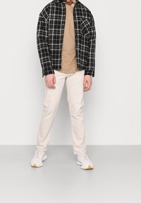 Camisa de franela a cuadros en blanco y negro sobre una camiseta beige, combinada con pantalones beige claros y zapatillas blancas con detalles en beige.