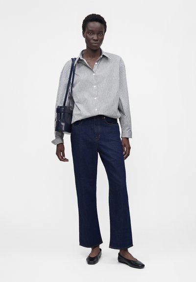 Γυναίκα που φοράει ριγέ πουκάμισο με κουμπιά, σκούρα τζιν με ίσια γραμμή, μαύρες μπαλαρίνες και κρατάει μια σκούρα μπλε τσάντα ώμου.