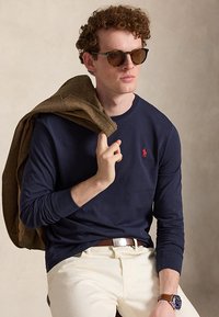 Mann mit lockigem Haar, der eine Sonnenbrille trägt, ein marineblaues Langarmshirt mit rotem Logo, weiße Hose, brauner Gürtel, Uhr, hält eine braune Jacke über der Schulter.