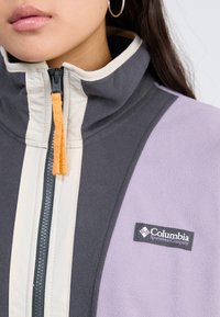 Fleece jacket in gray, lavender, and cream with a high collar, orange zipper pull, and Columbia Sportswear logo on the chest.