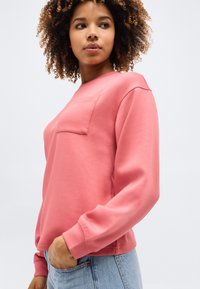 Γυναίκα με σγουρά μαλλιά που φοράει ροζ μακρυμάνικη μπλούζα με τσέπη στο στήθος, στέκεται με το ένα χέρι στην τσέπη ανοιχτόχρωμου τζιν.