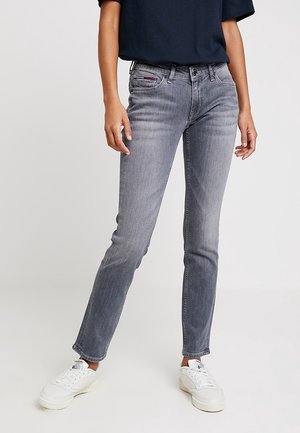 Personne portant un jean slim gris, un haut à manches courtes noir et des baskets blanches, debout devant un fond uni.