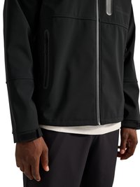 Zwart jasje met verticale ritssluitingzakken en verstelbare polssluitingen, gedragen over een wit overhemd en zwarte broek.